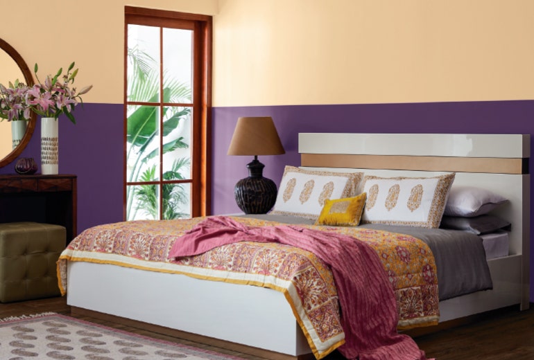 A bedroom with valley flower and purple diva shade walls, a bed by a wooden window, a bedside lamp, and a dressing table with a flower vase on it